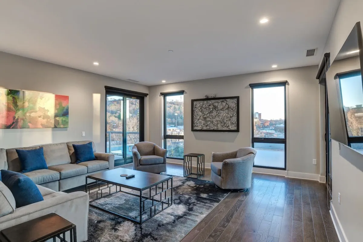 A spacious open-concept living room with a comfortable sofa, striking wall art, and a coffee table. Floor-to-ceiling windows invite natural light and offer sweeping views of the city and mountains beyond. The room is set up for both lively gatherings and peaceful relaxation, with contemporary decor and upscale furnishings.