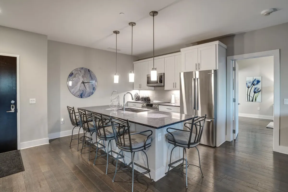 A bright and inviting kitchen featuring a large island with bar stools, modern appliances, and a wall clock. The open layout and seating area suggest a space perfect for gathering before heading out or sharing a casual meal.