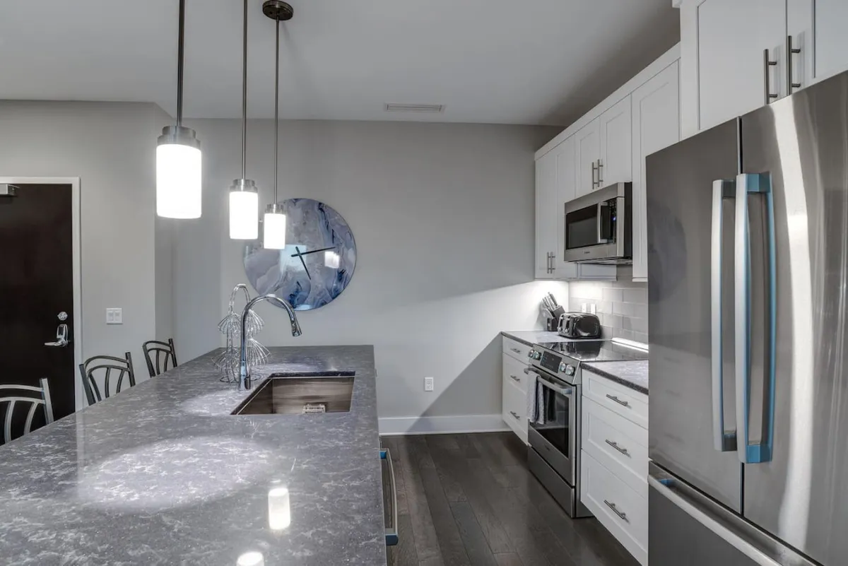 A modern kitchen area with a central island countertop, sleek stainless steel appliances, generous light fixtures, and vibrant blue wall art. The inviting space is perfect for casual dining, meal preparation, or enjoying a coffee break.