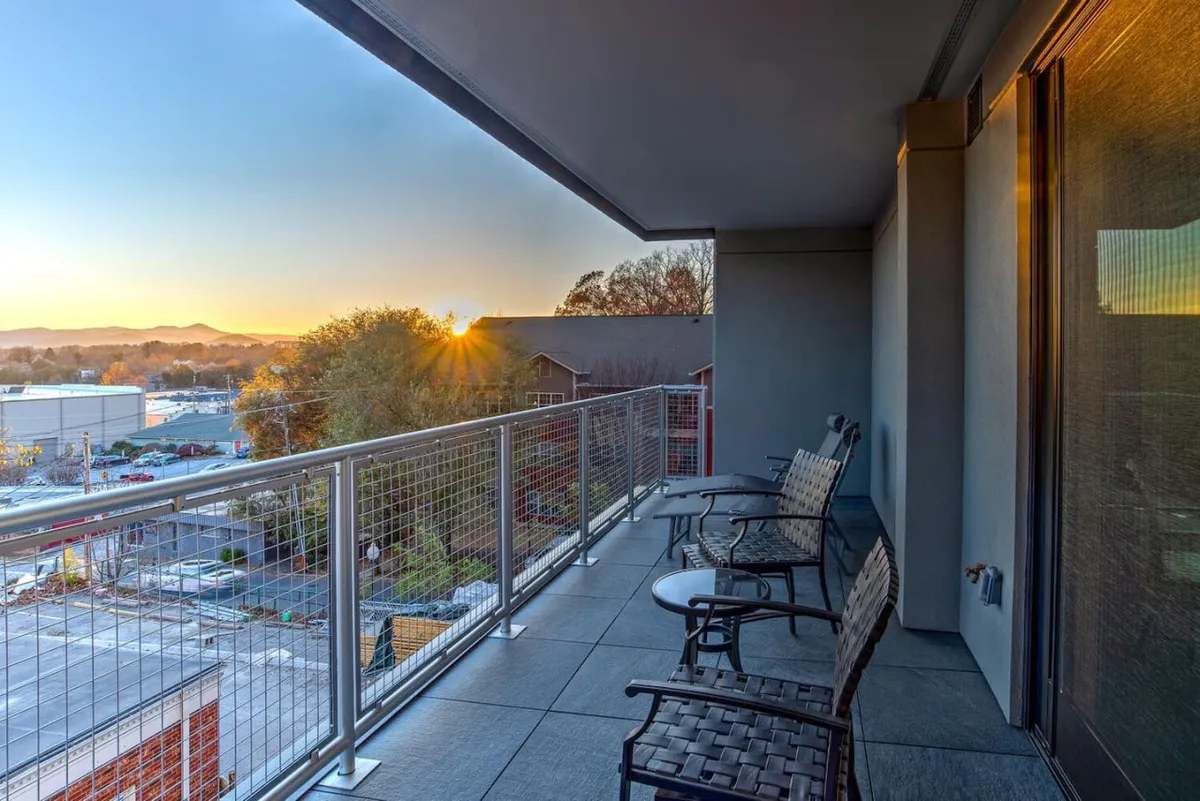 The photo features a cozy balcony setup with chairs arranged to face a stunning sunset view. The balcony railing frames the scene, underscoring the inviting outdoor space for morning coffee or evening relaxation above the city's bustle.