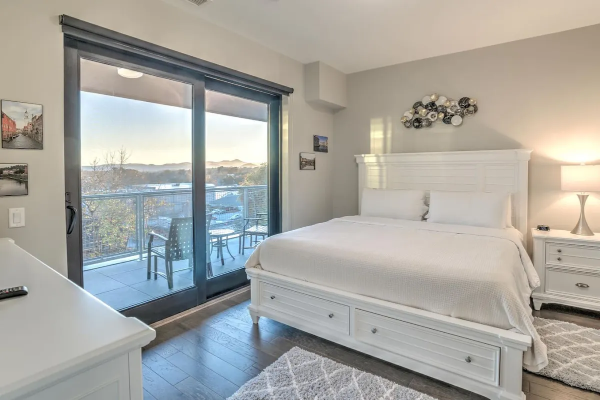This serene bedroom scene highlights a plush bed with luxurious bedding, stylish decorative wall art, and a nightstand with a lamp. Most importantly, the room opens directly onto a private balcony, offering a sweeping view of the Blue Ridge Mountains. The tranquil mood is perfect for relaxation, with natural light pouring in and a direct connection to the outdoors.