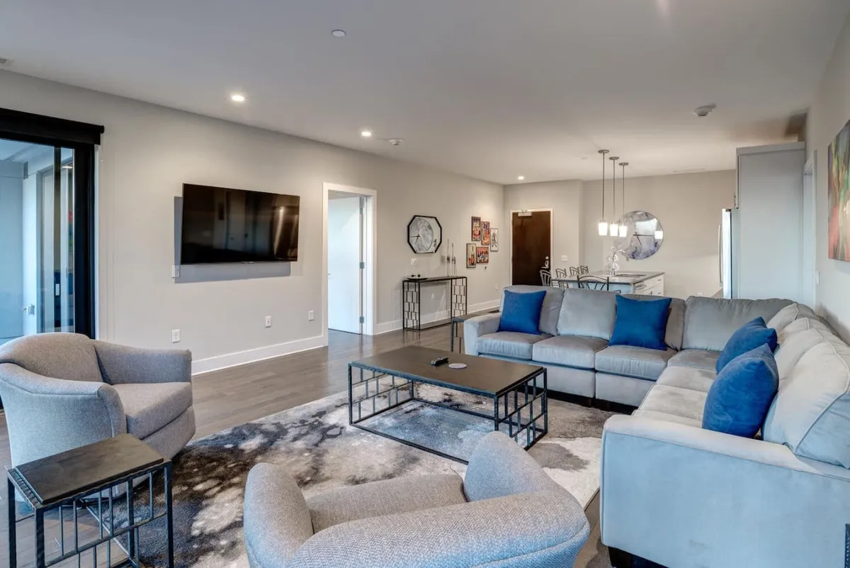 A spacious modern living room featuring a large sectional sofa with blue cushions, a coffee table, wall-mounted TV, and decorative wall art. The setting is comfortable and inviting, suggestive of a space where guests can relax after exploring the city.