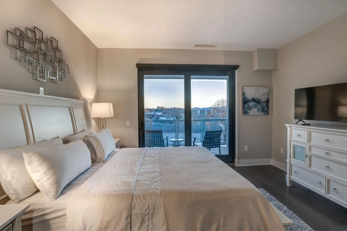 A bright bedroom with a large bed faces glass balcony doors, offering a direct view outside. Sunlight filters into the room, and beyond the doors lies a private balcony that hints at scenic vistas. The decor is peaceful, and the artwork and TV add touches of comfort. The connection to the outdoors is clear, inviting guests to start their day with beautiful views.