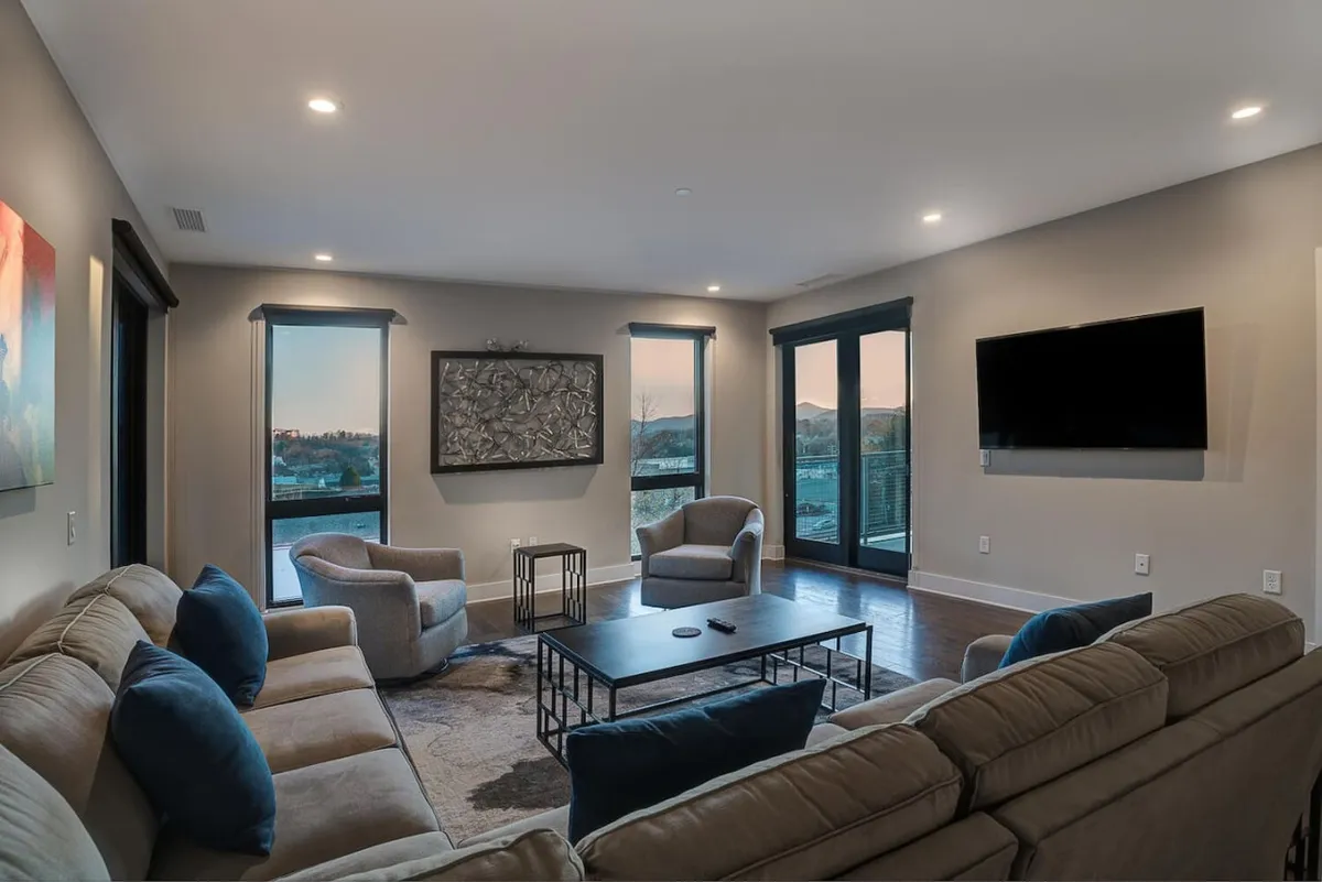 The image showcases a cozy, modern living room with a plush sofa, sleek coffee table, and large windows that frame sweeping sunset views of the Blue Ridge Mountains. The space is bathed in warm light, inviting guests to relax, watch TV, and share memorable moments together.