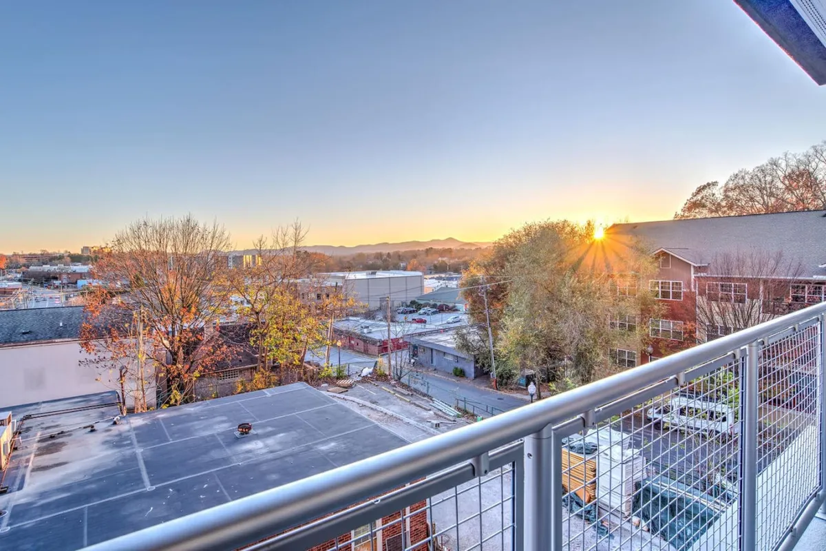 This image captures a sweeping view from the condo's balcony, showcasing a sunset over the mountains, the balcony railing, and the cityscape of downtown Asheville below. The warm evening light and vantage point evoke a sense of relaxation above the lively streets.